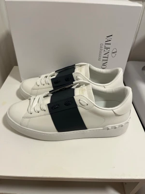 Valentino Garavani Open skor - Säljer ett par Valentino Garavani Open skor i vitt skinn med marinblått band över snörningen. Snygg och stilren design med rund tå, platt sula och ikoniska nitar på hälen. Perfekt för dig som gillar lyxiga och trendiga skor. Väldigt bra skick och är storlek 43