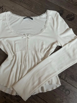 Vit långärmad blus med spets från Gina Tricot - Supersöt vit blus från Gina Tricot med lång ärm, spetsdetaljer runt halsringningen och framtill, samt en liten rosett vid urringningen. Blusen har en feminin, lätt rynkad midja och mjukt, stretchigt material som sitter bekvämt. Perfekt för en romantisk look.