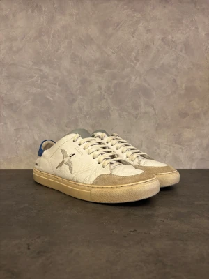 Axel Arigato Clean 90 - Snygga vita sneakers från Axel Arigato med blå häl och snygga Beebird designen. Skorna har snörning och en fågelbrodering på sidan. Tillverkade i skinn med platt sula och rund tå. Perfekta för dig som gillar stilrena och moderna sneakers. I använt skick och slitage efter användning finns på skorna, men inget som är sönder som hindrar användningen av skorna! Går att göras finare.