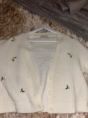 Vit blommig kofta från LCW Casual - Superfin vit stickad kofta från LCW Casual i storlek S. Koftan har broderade små blommor i gult och grönt, tre knappar framtill och ribbade muddar. Perfekt att slänga över en topp för en söt och chill look.