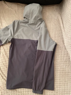Grå vindjacka från Under Armour - Säljer en snygg tvåfärgad vindjacka från Under Armour i ljusgrått och mörkgrått. Jackan har huva, hel dragkedja framtill, bröstficka med dragkedja och logga på bröstet. Perfekt för blåsiga dagar och outdoor-aktiviteter.