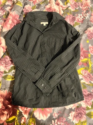 Svart randig skjorta med krage - Snygg svart skjorta med vita tunna ränder och klassisk krage. Skjortan har långa ärmar med knappar vid manschetterna och en rak passform. Perfekt att styla till jeans eller kjol för en clean look.