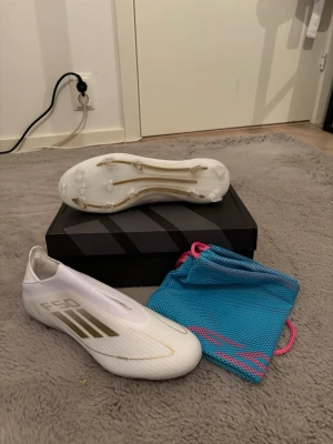 Adidas F50 vita fotbollsskor - Säljer ett par Adidas F50 fotbollsskor i vitt med guldiga detaljer och strömlinjeformad design. Skorna har platt sula med dobbar och är tillverkade i syntetmaterial för lätthet och snabbhet på planen. Kommer med en blå meshpåse med rosa snören.