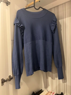 Blå stickad tröja med volang från Vila - Köpt för 400kr. Säljer en blå stickad tröja från Vila i storlek L. Tröjan har ribbad struktur, rund hals och långa ärmar med snygga volangdetaljer på axlarna. Perfekt för dig som gillar en enkel men ändå cool look.