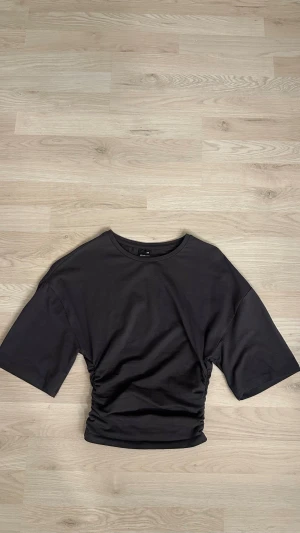 Mörkgrå croppad t-shirt från Gina Tricot - Mörkgrå t-shirt från Gina Tricot i storlek XXS. Modellen är croppad med snygg rynkning på sidorna och har korta men lite vidare ärmar. Super söt topp att matcha med enkla jeans. Knappast använd och är i jättebra skick. 