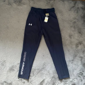 Mörkblå träningsbyxor Under Armour - Snygga mörkblå träningsbyxor från Under Armour med reflekterande logga på benet och elastisk midja med snörning. Byxorna har smal passform, dragkedja vid benslut. Storlek L men passar M. 