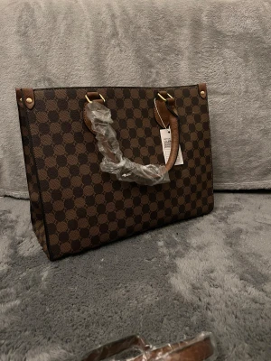 Brun rutig handväska med axelrem - Snygg handväska i brunt med rutigt mönster och guldfärgade detaljer. Väskan har både handtag och en avtagbar axelrem, vilket gör den enkel att bära på olika sätt. Materialet är skinnimitation och insidan är rymlig för dina grejer. Perfekt för dig som gillar klassisk stil med modern twist.