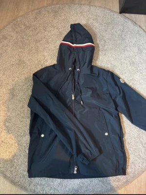 Mörkblå vindjacka från Moncler - Snygg mörkblå vindjacka från Moncler med huva och cool rödvit detalj på huvan. Jackan har dragkedja, snörning och två fickor med tryckknappar. Perfekt för dig som gillar stilrena och sportiga plagg. Materialet är lätt och känns skönt mot kroppen.