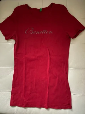 Röd kortärmad t-shirt från Benetton - Snygg röd t-shirt från Benetton med silverfärgat logotryck på bröstet. Modellen är klassisk med rund hals och korta ärmar. Materialet känns mjukt och skönt, 100% bomull. Hittade ingen storlek i tröjan, men skulle säga att den är S, kanske M