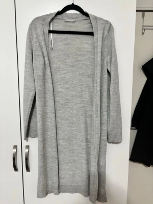 Lång grå kofta i mjukt material - Säljer en lång och skön grå kofta med ribbade detaljer vid krage och ärmslut. Koftan är öppen framtill och har långärmade ärmar, perfekt att slänga över en t-shirt eller topp. Enkel och stilren design som passar till många outfits.