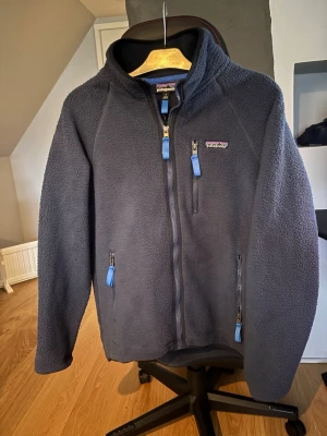 Patagonia fleecejacka  - Mysig mörkgrå fleecejacka från Patagonia med huva och tre fickor med blå dragkedjor. Jackan har lång ärm och är perfekt för kyliga dagar. Snygg, enkel design med broderad logga på bröstet och mjukt material som håller värmen. Skick 9/10. Hör av er vid frågor 