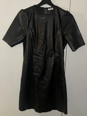 Svart kort klänning från Noisy May - Snygg svart klänning från Noisy May i storlek M. Klänningen är gjord i syntetmaterial med skinnlook, har rund halsringning och korta ärmar. Modellen är rak och slutar ovanför knäna, med dragkedja i ryggen för enkel påklädning.