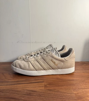 Beige Adidas Gazelle sneakers mocka - Säljer ett par stilrena Adidas Gazelle sneakers i beige mocka med vita detaljer och klassiska tre ränder på sidan. Skorna har rund tå, platt sula och snörning framtill. Perfekt för dig som gillar retrovibe och enkel design.