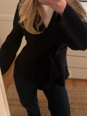 Mörkblå omlottkofta med knytband - Säljer en mörkblå omlottkofta med knytband i midjan. Koftan har långärmade ärmar och en stilren, lite loose passform. Perfekt att slänga på över en t-shirt eller linne för en chill och snygg look. Materialet känns mjukt och bekvämt mot huden.