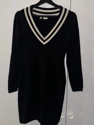 Svart stickad klänning Bodyflirt - Svart stickad klänning från Bodyflirt med vit-randig ribbad v-ringad krage. Klänningen har lång ärm och är kort i modellen, perfekt för dig som gillar sportig och stilren look. Materialet är mjukt och stretchigt, vilket gör den bekväm att bära.