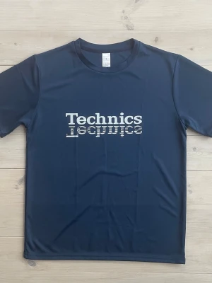 Deejay-T-shirt Technics - Snygg marinblå t-shirt från Technics med vit print på bröstet. T-shirten är gjord i mjuk polyester och har en klassisk rund hals och korta ärmar. Trycket har en cool grafisk effekt med ränder i bokstäverna. Perfekt för dig som gillar streetwear och musik. Den är ny och oanvänd. Den var för stor för mig. På etiketten står det S men det är Medium.