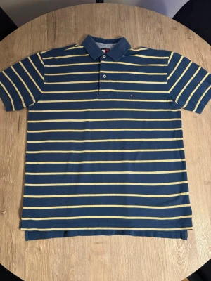 Pikétröja från Tommy Hilfiger - Säljer en klassisk pikétröja från Tommy Hilfiger i marinblått med gula horisontella ränder. Toppen har krage, korta ärmar och två knappar framtill. Materialet är mjuk bomull och den har en liten logga broderad på bröstet.