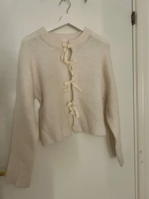Beige stickad kofta med knyt från Gina Tricot - Superfin beige stickad kofta från Gina Tricot med tre dekorativa knytband framtill. Koftan har långärmade ärmar och en mjuk, ribbad struktur som ger en mysig känsla. Perfekt att slänga på sig när det är lite kyligt ute.