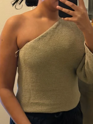 Beige stickad oneshoulder topp - Säljer en beige stickad topp med oneshoulder-design och lång ärm på ena sidan. Toppen har en avslappnad passform och är perfekt för dig som gillar en unik och trendig look. Materialet är mjukt och ger en skön känsla mot huden.