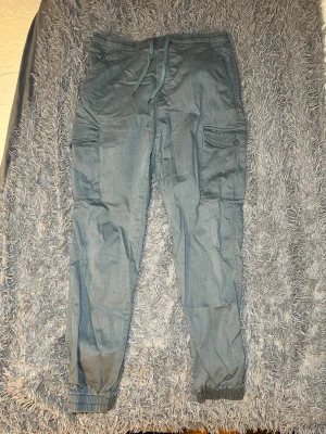 VAILENT Nico Cargobyxa, ljus blå/grå - Säljer ett par blå cargopants från Vailent i storlek L. Byxorna har elastisk midja med snörning, flera fickor med lock och knapp, samt resår vid bensluten. Materialet är 62% bomull, 32% polyester, 4% viskos och 2% elastan för skön passform. Nypris 599kr