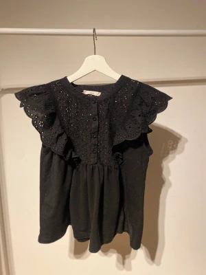 Svart blus med spets och volang från MNG - Supersöt svart blus från MNG med broderade blommor och spetsdetaljer på axlarna. Blusen har volang vid axlarna, knappar framtill och är ärmlös. Perfekt att styla med jeans eller kjol för en trendig look.