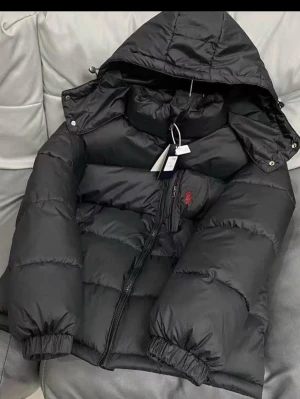 Svart pufferjacka med huva - Säljer en svart pufferjacka från Polo Ralph Lauren med huva och dragkedja framtill. Jackan har ett broderat rött logomärke på bröstet och elastiska muddar vid ärmslut. Perfekt för kalla dagar och har en riktigt schysst oversized look.