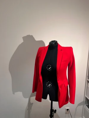 Röd kavaj med klassisk passform - Snygg röd kavaj med figurnära siluett och klassisk krage. Jackan har två knappar framtill, fickor med lock och en bröstficka. Materialet är slätt och känns som polyester eller bomull. Perfekt för dig som vill sticka ut med färg.