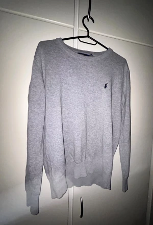 Grå tröja Polo Ralph Lauren - Snygg grå tröja från Polo Ralph Lauren med klassisk rund hals och diskret broderad logga på bröstet. Tröjan har långärmad passform och ribbade muddar vid ärmslut och nederkant. Perfekt för dig som gillar stilrena och tidlösa plagg.