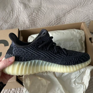 Adidas Yeezy Boost 350 V2 Carbon - Säljer ett par Adidas Yeezy Boost 350 V2 Carbon med svart och grå stickad ovandel, snörning och en ljus, ribbad Boost-sula. Skorna har en modern och chunky siluett med rund tå och platt sula. Perfekt för dig som gillar streetwear och vill sticka ut.