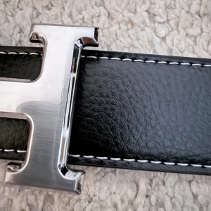 Svart skinnbälte med H-spänne - Snyggt svart bälte i skinn med ett stort, silvrigt H-format spänne. Bältet har vita sömmar längs kanterna som ger en schysst kontrast. Perfekt för att lyfta din outfit med en stilren detalj. Passar till jeans eller kostymbyxor.