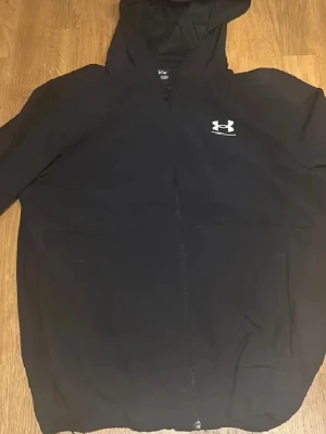 Svart vindjacka från Under Armour - Svart vindjacka från Under Armour med huva och hel dragkedja framtill. Jackan har ett stilrent vitt logotryck på bröstet och är tillverkad i lätt polyester med meshfoder. Perfekt för aktiva dagar och sportiga outfits.