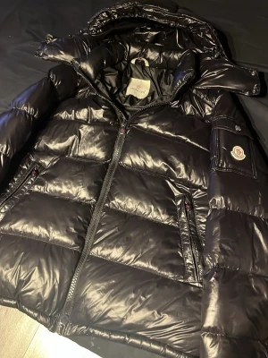 Svart Moncler pufferjacka med huva - Säljer en svart pufferjacka från Moncler med glansig finish och avtagbar huva. Jackan har dragkedja framtill, två sidofickor med dragkedja och Moncler-logga på ärmen med NFC tagg. Materialet är syntet och dun, perfekt för kalla dagar. Insidan har ett coolt serietidningsmönster och en liten ficka med dragkedja. Väldigt varm