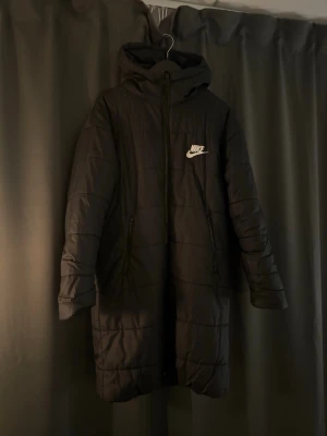 Svart lång pufferjacka från Nike - Svart lång pufferjacka från Nike med huva och dragkedja framtill. Jackan har vita Nike-loggor både på bröstet och ryggen, samt två sidofickor med dragkedja. Quiltad design och stilren look, perfekt för kalla dagar.
