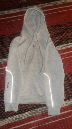 Nike NOCTA grå mjukisbyxor - Säljer ett par grå mjukisbyxor från Nike NOCTA med svarta loggor på benet och reflekterande detaljer längs sidorna. Byxorna har elastisk midja och muddar vid benslut, perfekt för chill eller streetwear. Materialet är mjukt och bekvämt, troligen bomull med inslag av polyester.