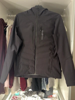 Svart vindjacka från Sail Racing - Svart vindjacka från Sail Racing med huva och flera dragkedjeförsedda fickor. Jackan har stilren design med logga på bröstet och detaljer i silver. Materialet är slitstark polyester och passformen är normal. Perfekt för blåsiga dagar och outdoor.