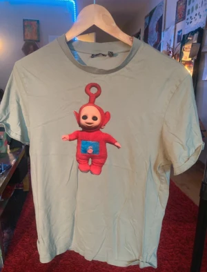 Grå t-shirt med unikt Teletubbies-tryck - Ljusgrå t-shirt med tryck av ett konstverk jag skapat. Handtryckt av mig :)   Perfekt för den som vill ha något helt unikt då det bara finns ett enda exemplar!  Själva nallen säljes också. 🥳🥳🥳