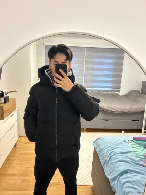 Svart pufferjacka från Superdry - Säljer en svart pufferjacka från Superdry med huva och dragkedja framtill. Jackan har en clean look med diskreta logodetaljer på ärmen och insidan. Perfekt för kalla dagar och riktigt skön att bära tack vare den vadderade designen.