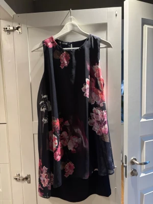 Svart blommig klänning Bodyflirt S - Svart kortklänning från Bodyflirt i storlek S med ett snyggt blommigt mönster i rosa och rött. Klänningen är ärmlös och har en luftig, lätt form med rund halsringning. Perfekt för dig som gillar färgstarka detaljer och en flowy siluett.