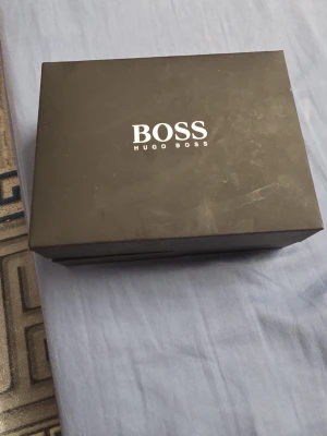 Hugo boss klocka  - Bilderna visar en Hugo Boss-klocka med svart läderarmband och ett svart flätat armband, samt en svart box med BOSS-logga. Ingen väst syns på bilderna. Priset kan diskuteras 
