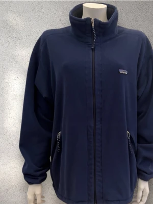 Fleecejacka från Patagonia - Mörkblå fleecejacka från Patagonia med hög krage och hel dragkedja framtill. Jackan har två sidofickor med dragkedja och ett litet broderat logomärke på bröstet. Materialet är mjuk fleece och modellen är loose fit.                        PS: Lätt slitage på insidan. Syns inte vid användning och påverkar inte funktionen se bilder