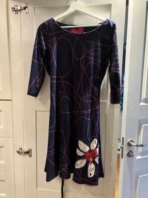 Mönstrad klänning från Desigual - Säljer en cool marinblå klänning från Desigual med rosa slingrande mönster och en stor vit blomma med rött hjärta på nederdelen. Klänningen har trekvartsärm och är i mjukt material, perfekt för dig som gillar färg och unika detaljer.