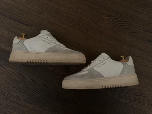 Vita sneakers från Axel Arigato - Snygga vita sneakers från Axel Arigato med beige detaljer och platt sula. Skorna har rund tå, snörning och är tillverkade i skinn och mocka. Perfekta för dig som gillar stilrena och moderna sneakers med en lyxig känsla.