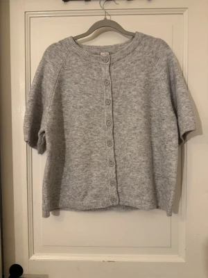 (Ny) Grå stickad kofta från Nelly - Mysig grå stickad kofta från Nelly med korta ärmar och rund hals. Koftan har knappar hela vägen framtill och ribbade detaljer vid hals, ärmslut och nederkant. 