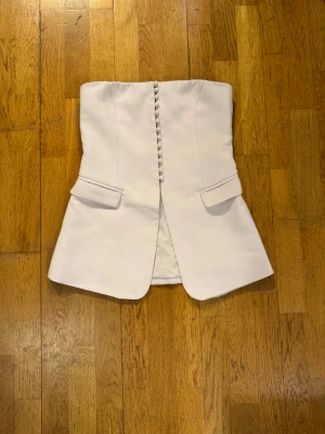 Vit korsettopp från boa med knappar S - Supersnygg vit korsettopp från boa i figurnära modell. Toppen är axelbandslös och har dekorativa klädda knappar framtill samt två fejkfickor. Materialet känns som en blandning av polyester och bomull, perfekt för att skapa en chic look.