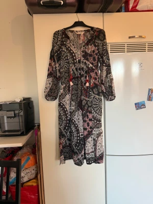 Odd Molly långklänning blommig - Säljer en långklänning från Odd Molly med blommigt och paisleyinspirerat mönster i svart, vit, röd och beige. Klänningen har långa ärmar, v-ringning med snörning och dekorativa tofsar framtill. Materialet är tunn bomull som känns skönt mot huden. Storlek 0 som enligt deras storleksguide motsvarar en XS. Mer en S kanske liten M,  Mycket fint skick, jag hittar inga synliga anmärkningar eller defekter.  