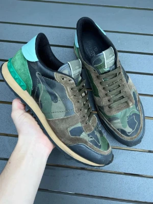 Valentino Rockrunner camo sneakers - Säljer ett par Valentino Rockrunner sneakers med camouflagemönster i grönt, skorna är i storlek 45, brunt och svart. Skorna har detaljer i mocka och skinn, samt en cool grön häl med nitar och ljusblå läderpanel. Platt sula och snörning framtill. Perfekt för dig som gillar streetstyle. Hör av er för minsta fundering,💨