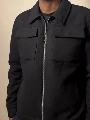 Svart overshirt med dragkedja - Svart overshirt med klassisk krage och två stora bröstfickor. Jackan har en rak passform och är tillverkad i ett tjockare material som känns lite ulligt. Stängs med en silverfärgad dragkedja framtill. Perfekt för dig som gillar stilrena och enkla plagg.