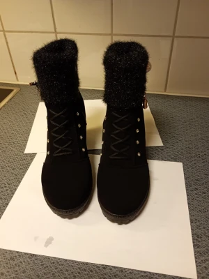 Svarta stövletter med pärldetaljstl 40 - Säljer ett par svarta stövletter med fluffig, glittrig kant upptill och snygga pärldetaljer på sidan. Skorna har snörning framtill, dragkedja på sidan och dekorativa nitar. Klacken är stabil och mellanhög, perfekt för dig som gillar att sticka ut.stl 40