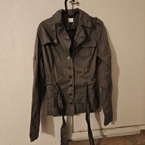 Grå tunn jacka från Oxmo med bälte - Snygg grå parkajacka från Oxmo med knappar framtill, flera fickor och bälte i midjan. Jackan har lång ärm och klassisk krage, perfekt för dig som gillar en clean och chill stil. .  Nytt med lappen kvar. Jag kan inte se några hål eller skador etc. Även om de är nytt.  Obs! Jag står inte för frakbolagets slarv, skador eller annat som händer efter att jag har postat.