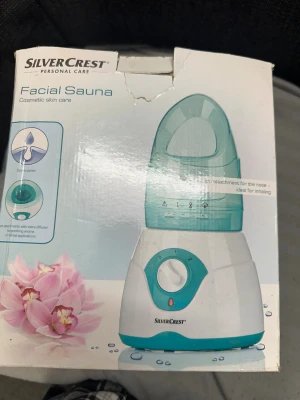SilverCrest ansiktssauna med tillbehör - Elektrisk ansiktssauna i vit och turkos plast från SilverCrest. Kommer med flera utbytbara munstycken och en liten mätkopp. Perfekt för att öppna porer och ge huden en fräsch känsla. Enkel att använda med vred för inställning av ångnivå. ( nytt skick använd bara 3 gånger bara kartongen är skadad)